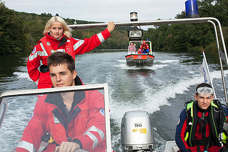 2 Einsatzkräfte der Wasserwacht und ein Taucher fahren in einem Boot, ein anderes Boot fährt in deren Kielwasser