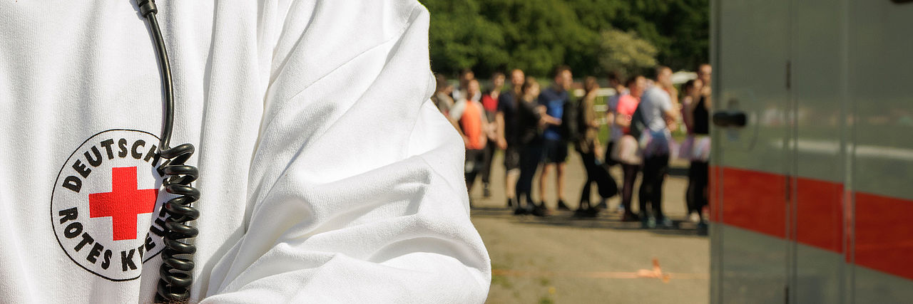 Ehrenamtlicher des DRK mit Funkgerät und weißem Pullover vor einer wartenden Menschenschlange