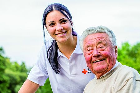 Pflegekraft steht hinter Senior, beide lächeln in die Kamera