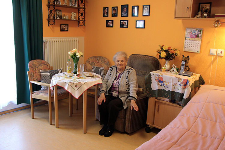 Eine Frau sitzt in ihrem Zimmer in unserer Einrichtung auf einem Sessel. Im Hintergrund sieht man Bilder und andere Möbel