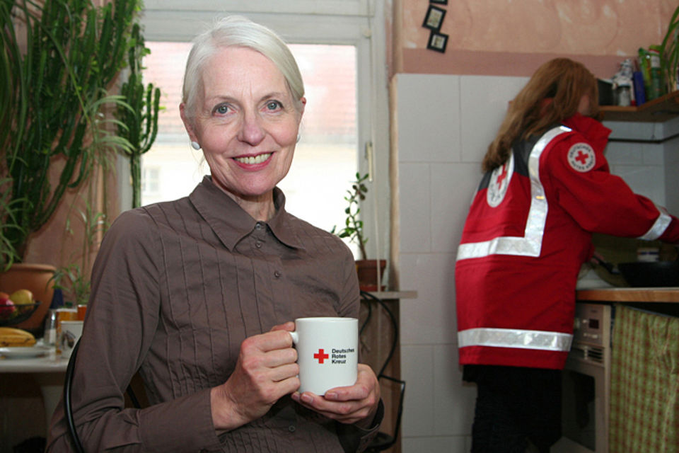 Eine ältere Frau mit einer DRK-Tasse in der Hand. Im Hintergrund ist eine Mitarbeiterin des DRK an der Küchenspüle.