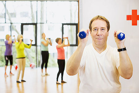 Ein Mann hebt mit jeder Hand eine Hantel. Im Hintergrund sieht man mehrere Personen, die Fitnessübungen machen