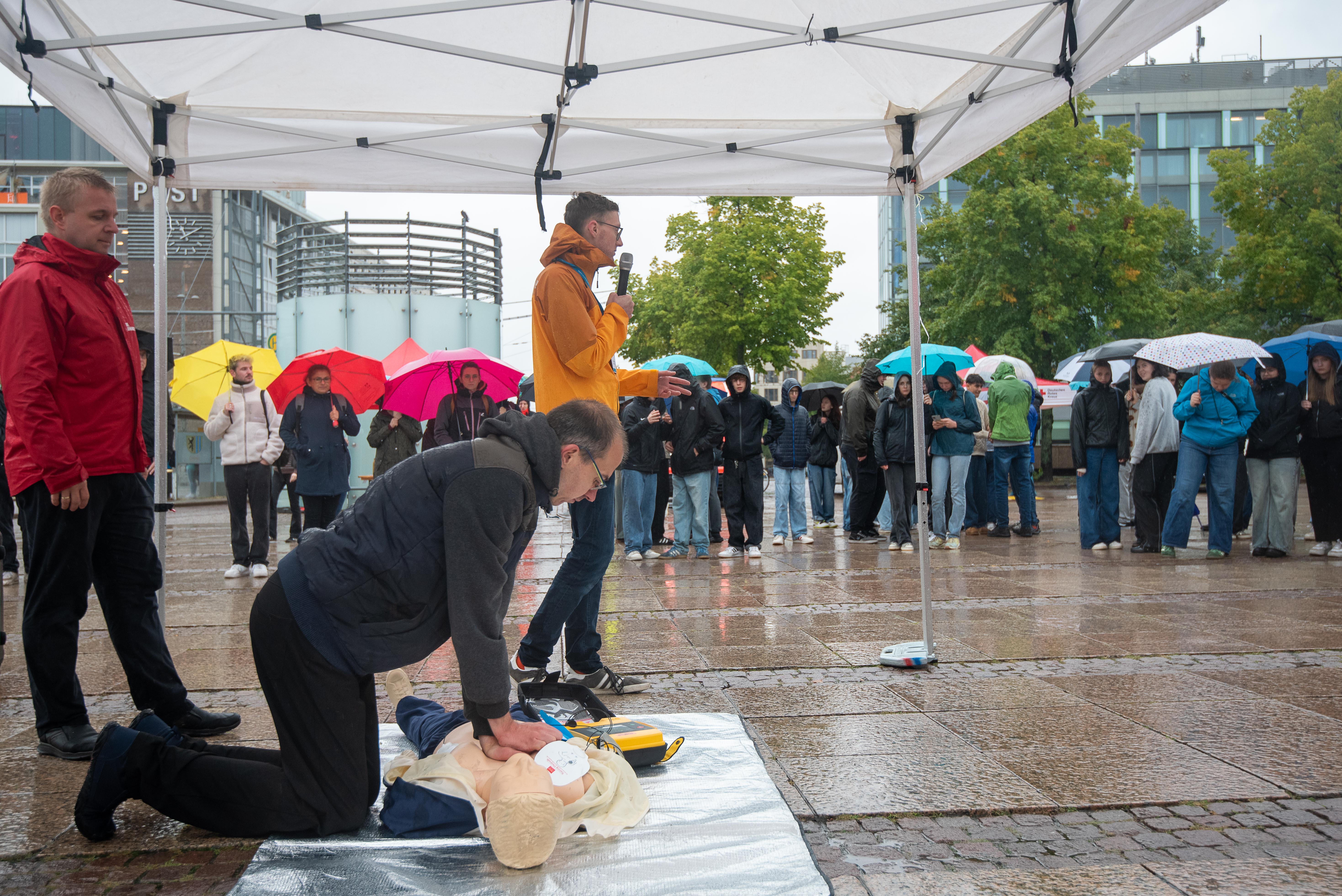 Mann führt Wiederbelebung an Puppe durch, ein anderer erklärt die Schritte den Umstehenden